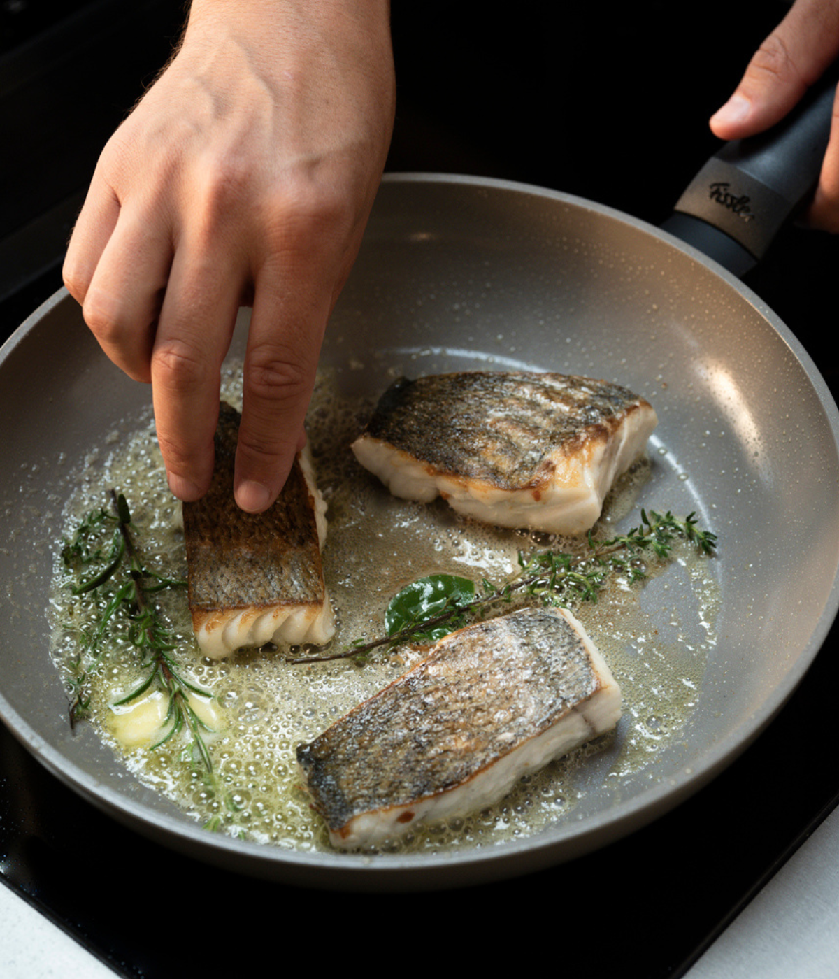 Three pieces of skin-on fish are expertly seared in a Fissler frying pan, surrounded by bubbling butter, fresh herbs, and garlic—demonstrating precision cooking with German-engineered quality cookware.