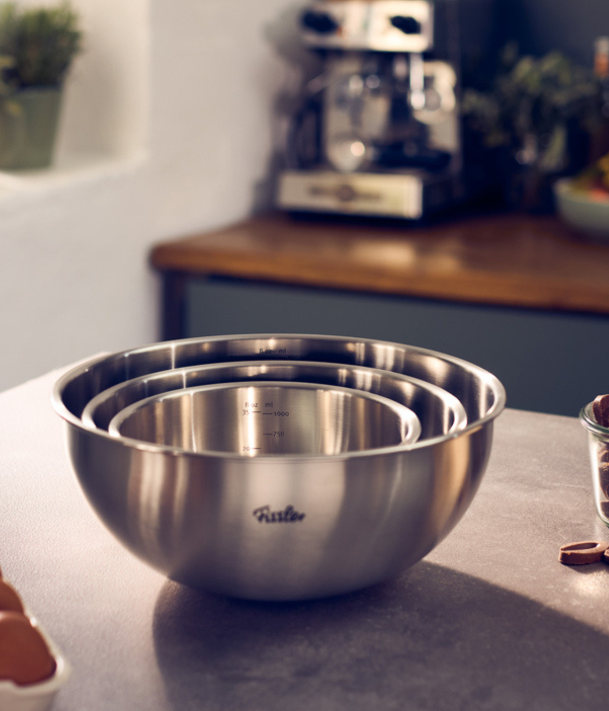 Three premium Fissler stainless steel mixing bowls, elegantly nested together, rest on a kitchen countertop. Precision-engineered with signature German craftsmanship, their polished finish reflects the brand’s timeless design. In the background, a coffee machine and potted plant highlight a sophisticated kitchen setting.