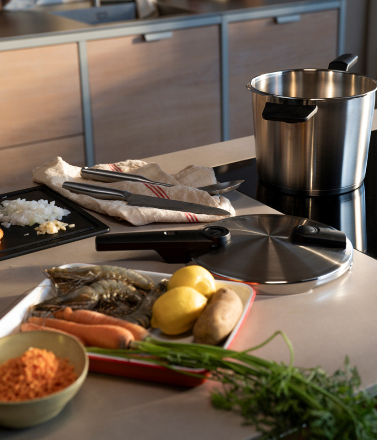 A kitchen countertop featuring a Fissler stainless steel pot with matching lid, surrounded by fresh carrots, lemons, potatoes, chopped onions, and a bowl of red lentils. High-quality Fissler cooking utensils are neatly arranged on a towel nearby, highlighting precision German engineering and timeless design.