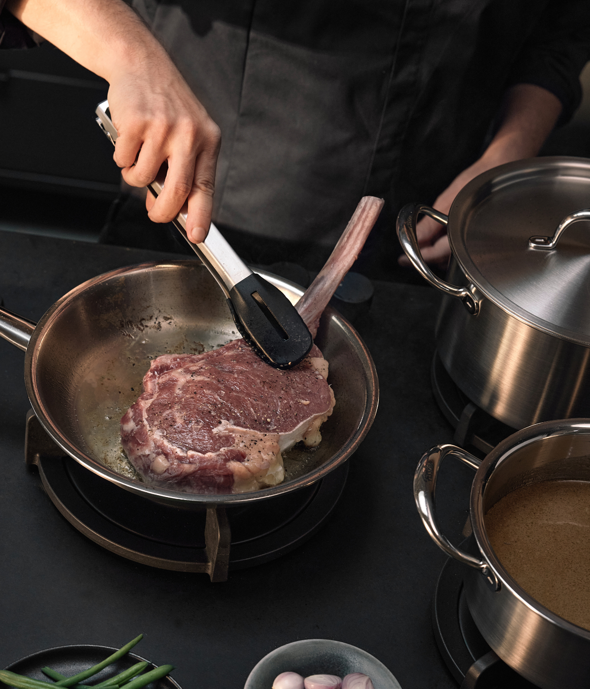 A person expertly sears a large bone-in steak in a premium Fissler stainless steel pan using tongs. Nearby, two Fissler saucepans rest on the stovetop, crafted with precision and durability, while fresh green beans and shallots are visible at the bottom of the frame, highlighting a kitchen scene defined by German engineering and timeless design.