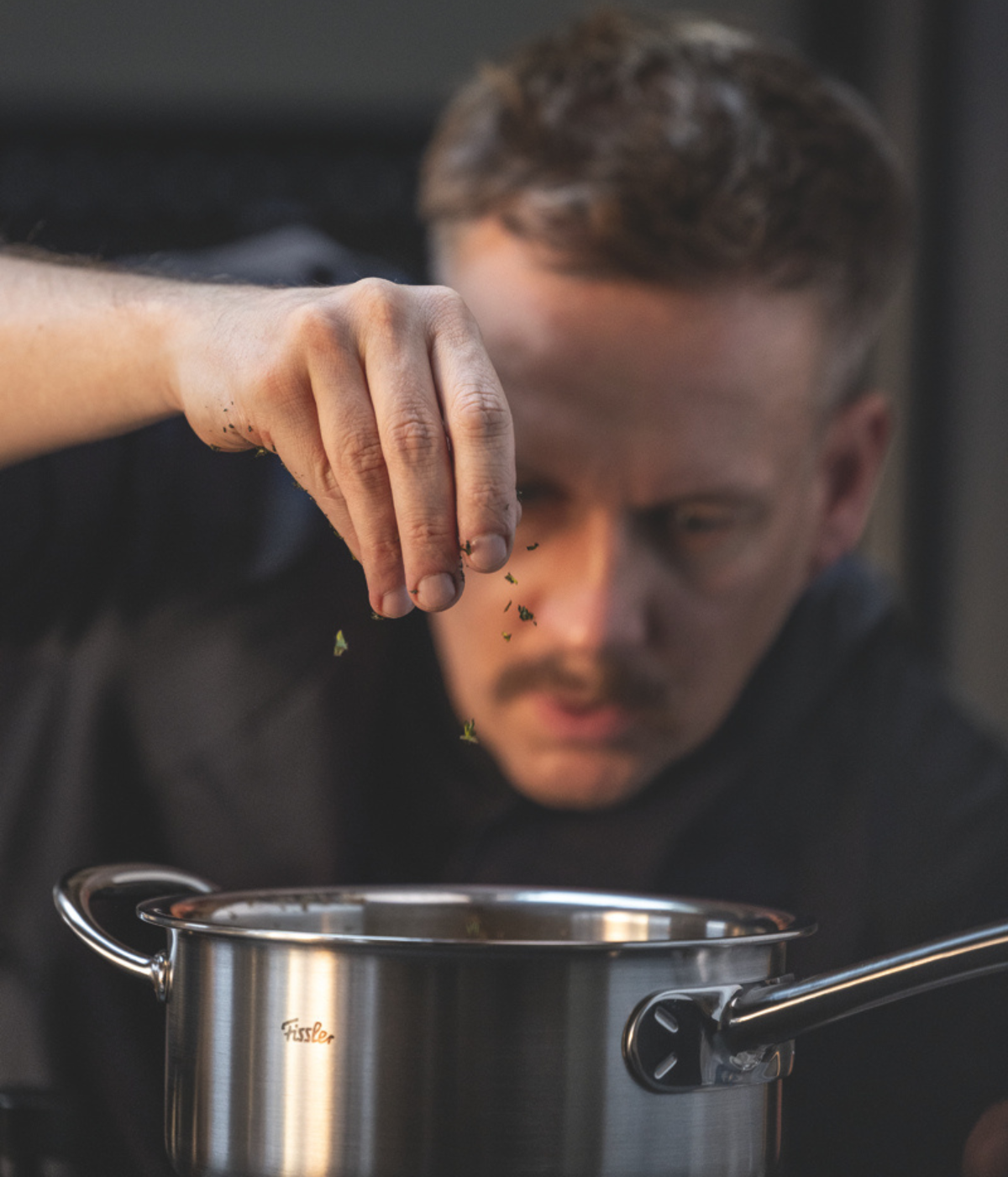 A chef adds fresh herbs to a Fissler stainless steel pot, showcasing precision and focus, with the pot’s sleek German engineering and timeless design evident in the kitchen scene.