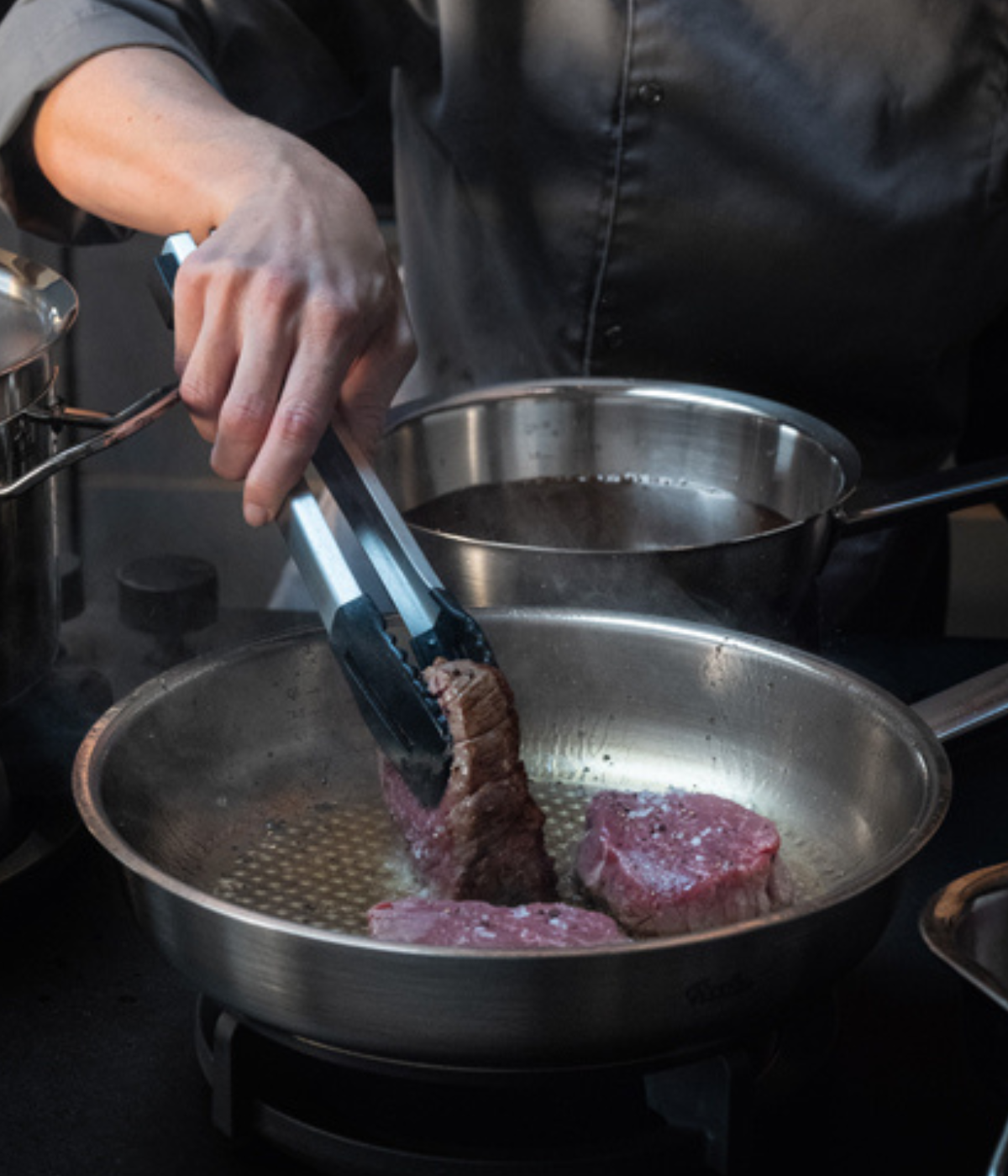 A chef in a dark jacket expertly sears steak pieces using tongs in a Fissler stainless steel pan on the stove, highlighting precision engineering and timeless German design.