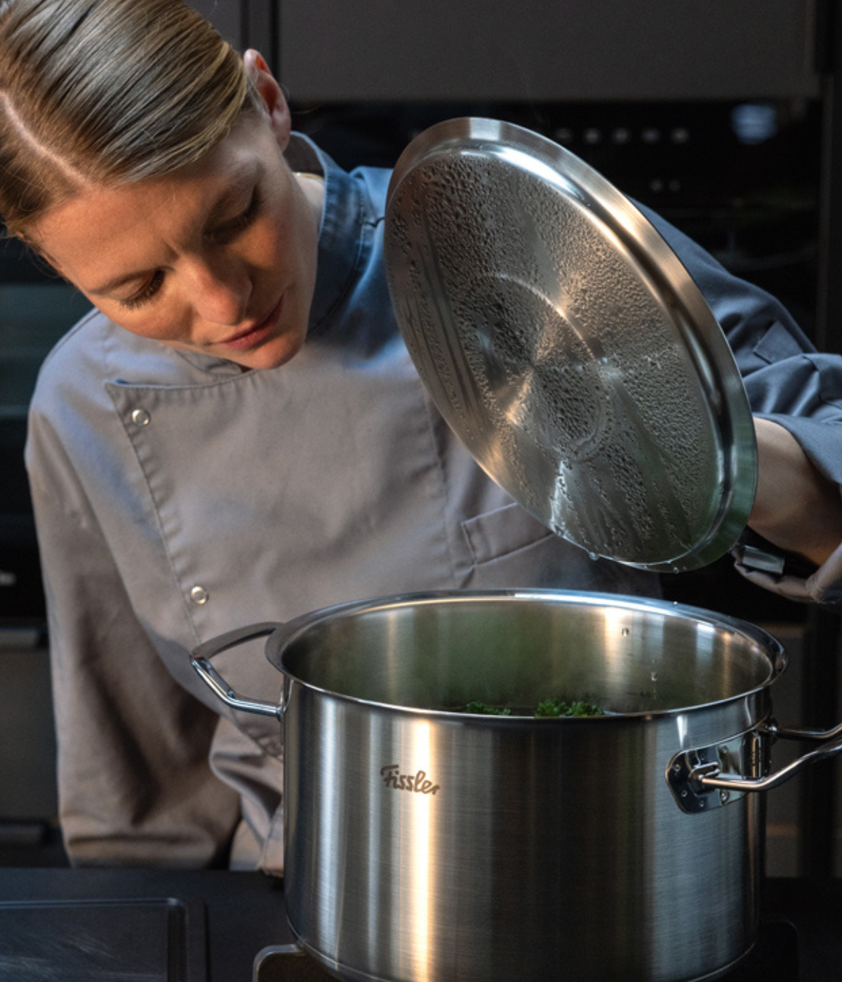 Ein Koch in grauer Uniform hebt den Deckel eines hochwertigen Fissler-Edelstahltopfs auf dem Herd an und präsentiert dabei präzise Technik und zeitloses deutsches Design, während er die Zutaten im Topf überprüft.