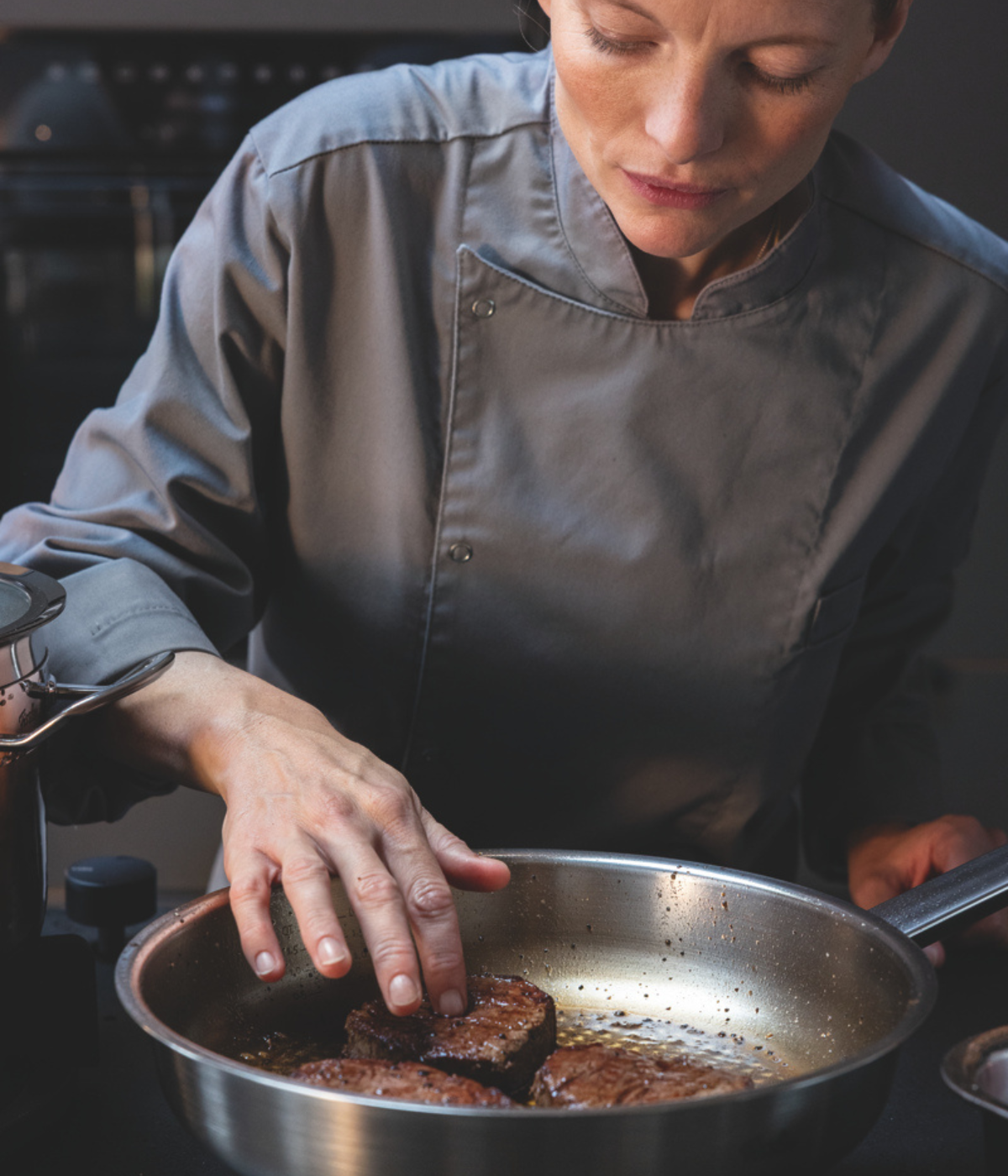 A chef in a grey uniform checks the doneness of two steaks sizzling in a Fissler stainless steel pan, showcasing precision and quality engineering “Made in Germany.“.