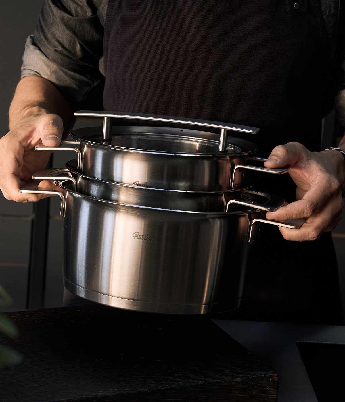 A person in a dark apron showcases two Fissler stainless steel pots with ergonomic handles, elegantly nested together, reflecting the brand’s precision engineering and timeless German design in a modern kitchen setting.