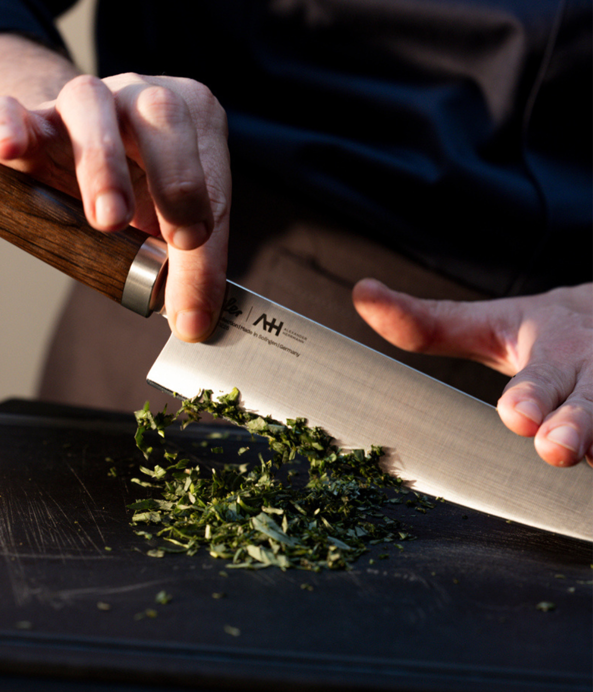 A person precisely chops fresh herbs on a sleek black cutting board using a premium Fissler kitchen knife with an elegantly crafted wooden handle, reflecting the brand’s commitment to German engineering, durability, and timeless design.