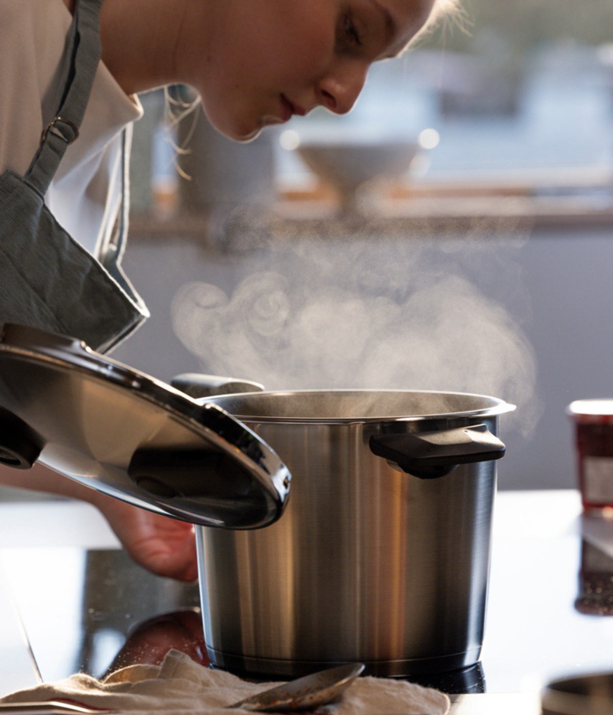 In a sunlit kitchen, a person in an apron carefully lifts the lid from a steaming Fissler stainless steel pot, showcasing the brand’s signature precision engineering and timeless German design. Sunlight streams through the window, illuminating the high-quality cookware at the heart of Fissler’s heritage collection.