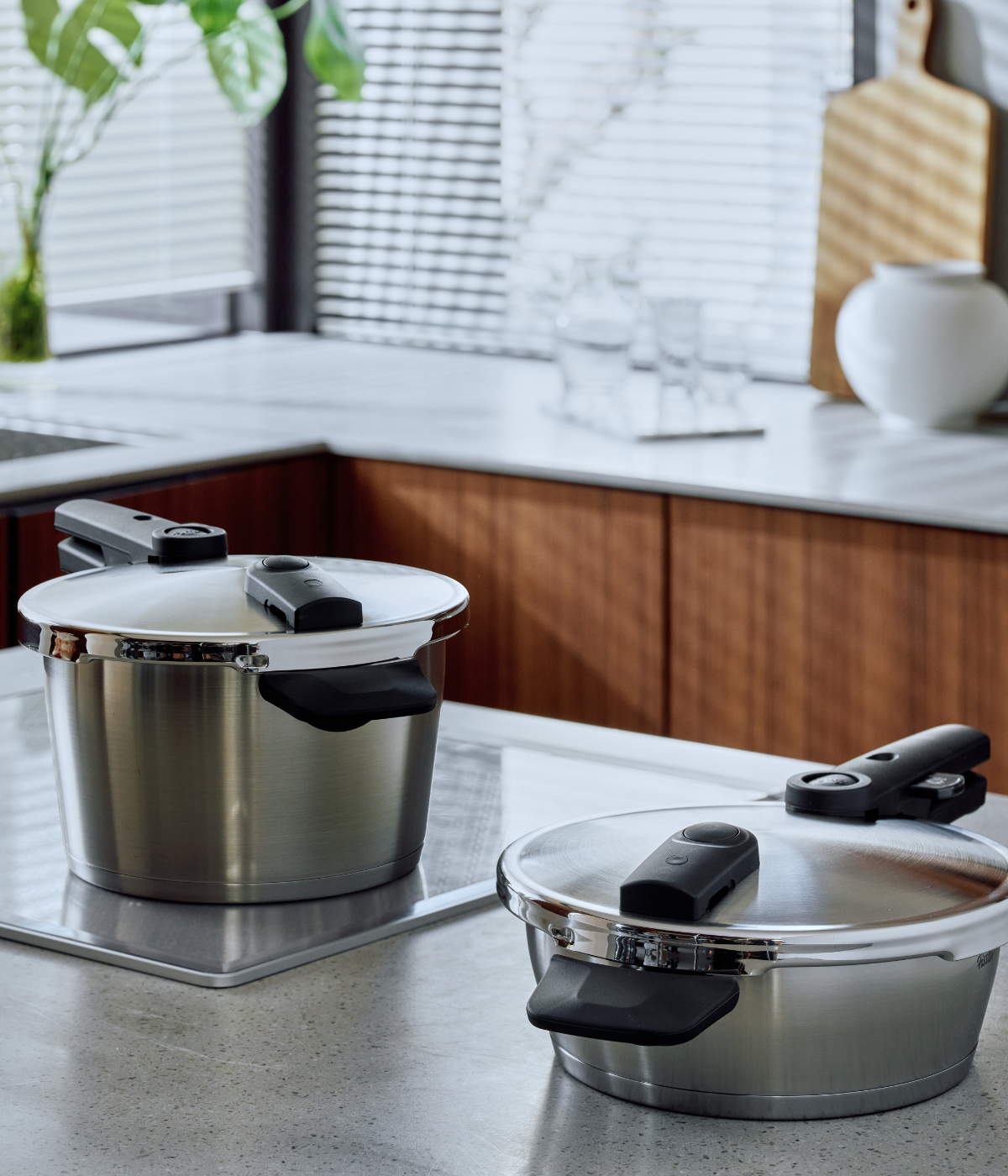 Two Fissler stainless steel pressure cookers with signature black handles rest on a modern kitchen countertop. Sunlight filters through window blinds, illuminating the white marble surfaces and highlighting the refined durability and precise German engineering of these high-quality cookware pieces. Elegant wooden cabinets serve as a backdrop, reflecting Fissler’s heritage of timeless design and innovation since 1845.