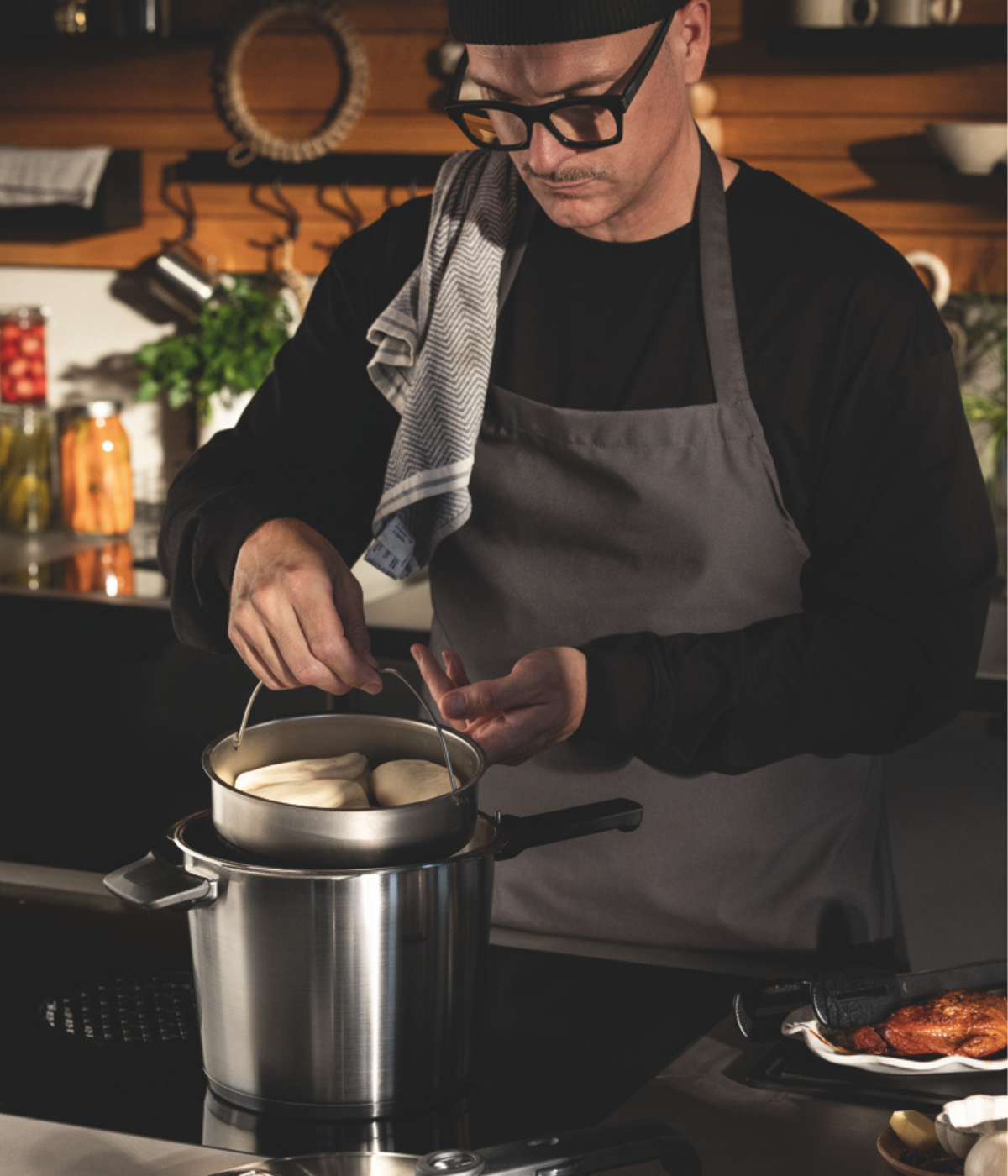 A man wearing glasses, a hat, and an apron expertly steams a tray of buns over a premium Fissler stainless steel pot in a well-appointed kitchen. Shelves lined with jars provide an inviting backdrop, highlighting the precision engineering and timeless design of Fissler cookware at the heart of the cooking process.