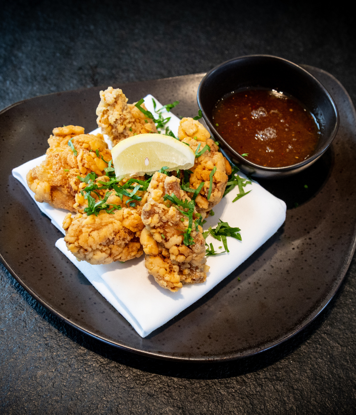 Crispy fried chicken pieces, garnished with fresh herbs and a lemon wedge, are elegantly presented on a sleek black plate atop a napkin, accompanied by a small bowl of dipping sauce. The refined plating highlights the quality cookware and timeless design that Fissler is renowned for—engineered for durability and precise results in every kitchen creation.
