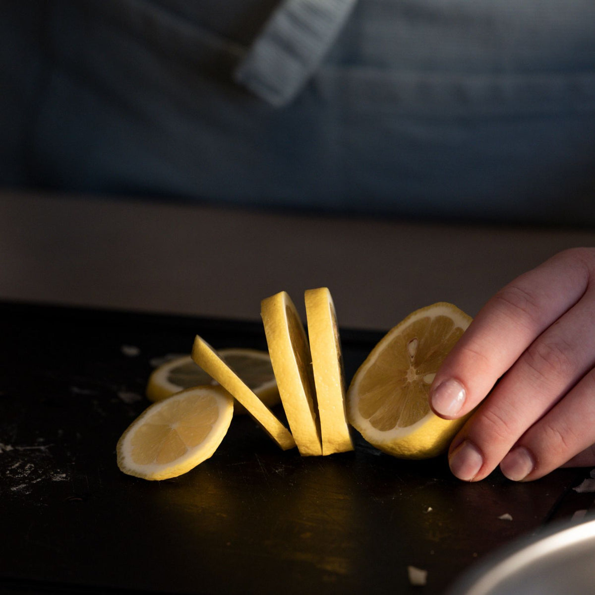 A hand prepares a lemon on a smooth black Fissler cutting board, with slices artfully arranged; the person wears a blue apron.