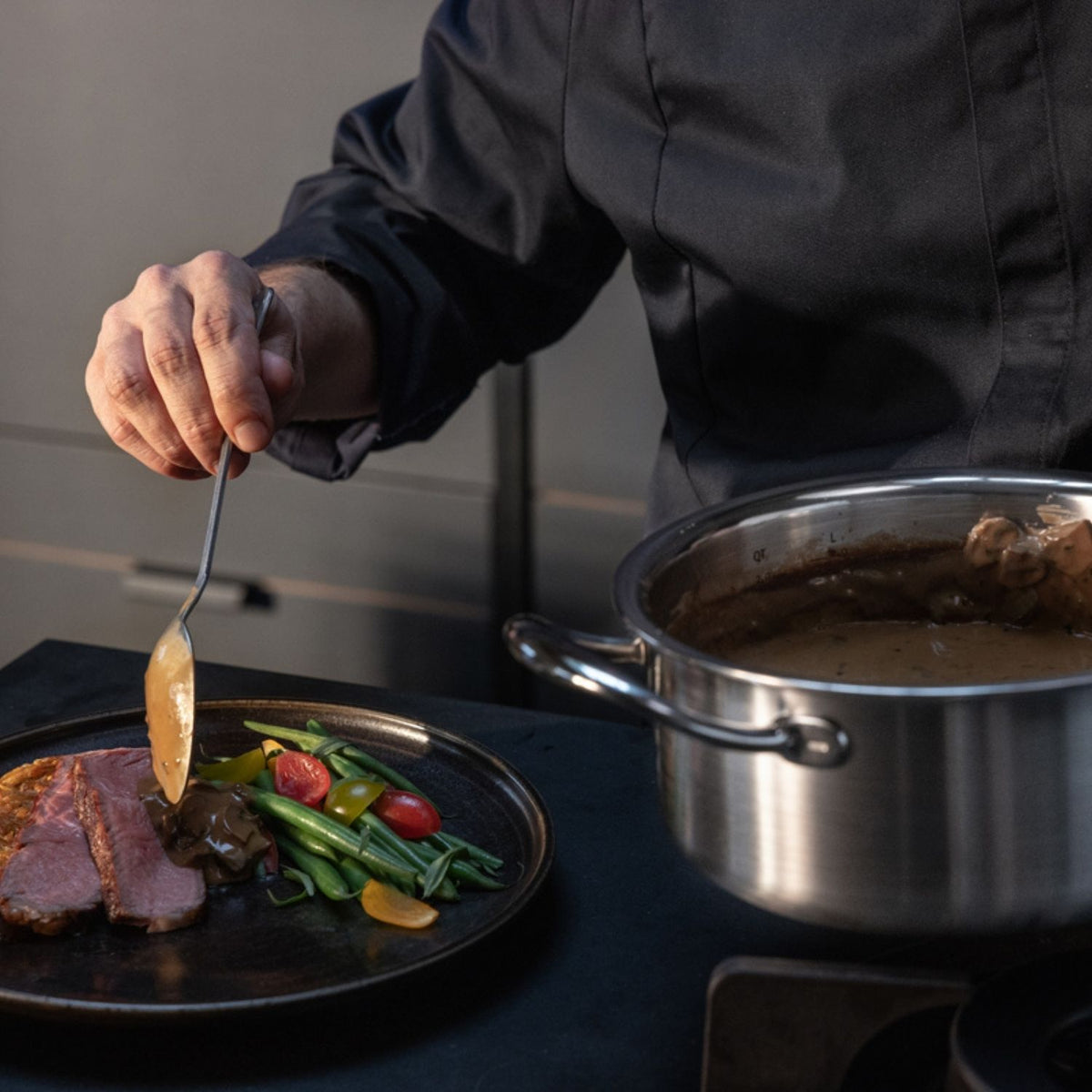 Ein Koch verwendet einen Fissler-Edelstahltopf, um Sauce über Fleisch, grüne Bohnen und Tomaten zu servieren und präsentiert damit deutsches Präzisionskochgeschirr.