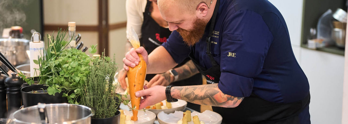 A chef in navy blue uses a piping bag to plate food next to Fissler cookware, with herbs and utensils on the counter.