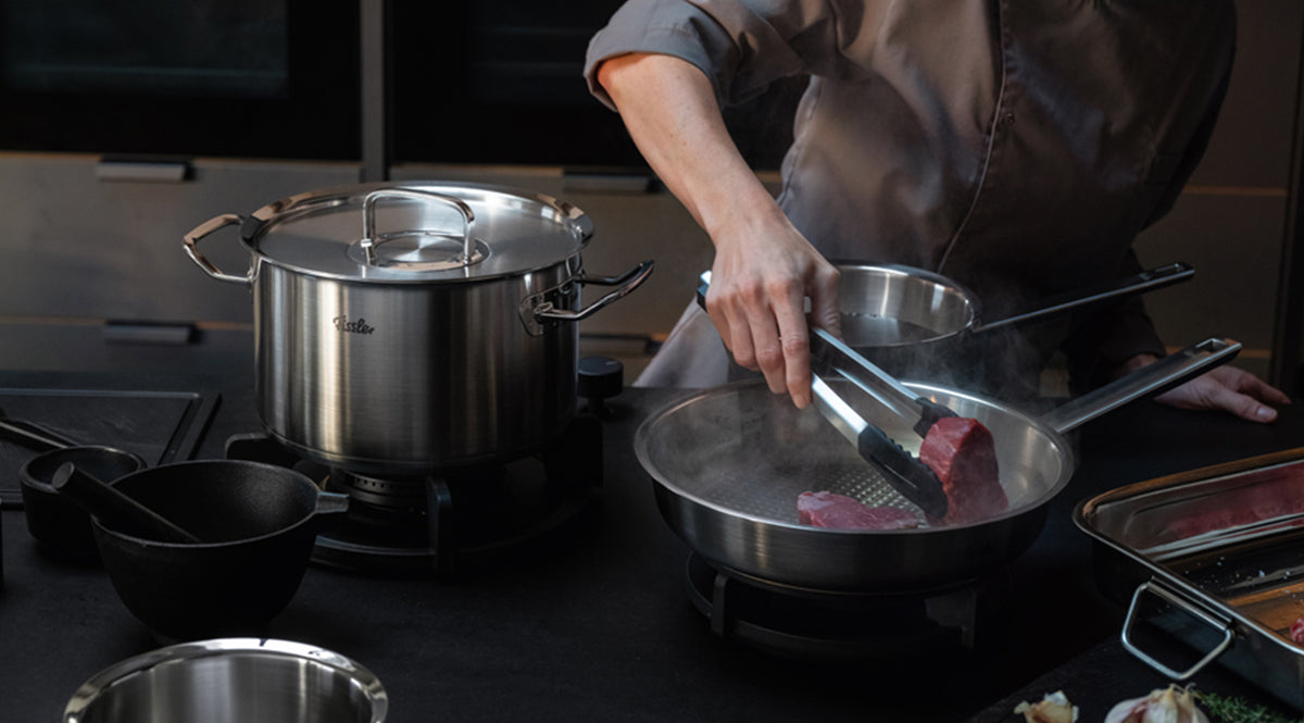 A chef in a professional jacket expertly sears a cut of meat in a Fissler stainless steel skillet using tongs. Beside them, a sizable Fissler pot sits on the stove, surrounded by premium kitchen utensils on an elegant dark countertop, showcasing the precision engineering and timeless design of Fissler cookware made in Germany.