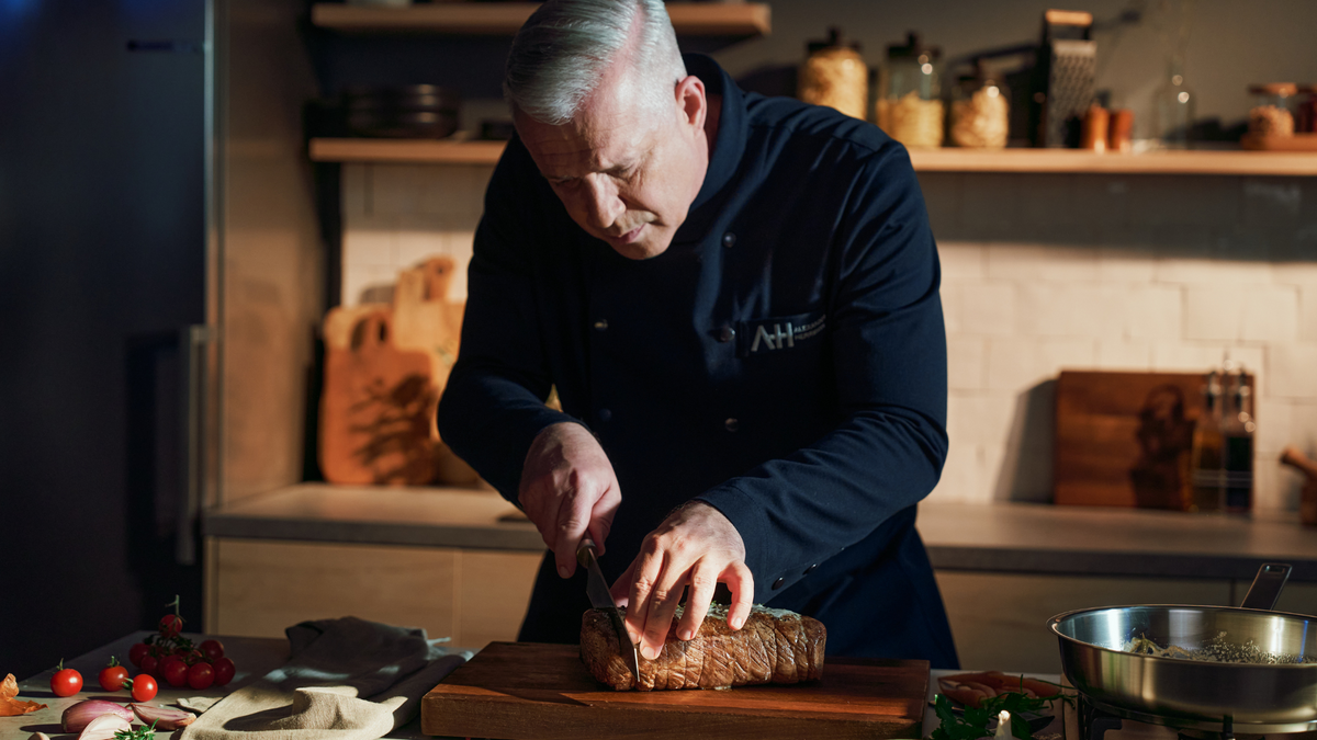 A chef with gray hair in a dark uniform slices roast on a wooden board in an elegant, modern kitchen with Fissler cookware.