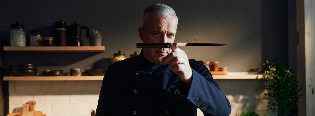 A chef in a modern kitchen inspects a chef's knife, surrounded by Fissler stainless steel cookware and premium kitchen appliances.