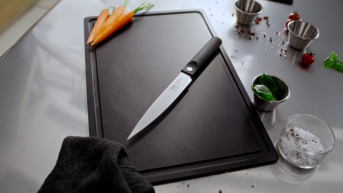 Das Fissler ProCut Fleischmesser (18 cm), gefertigt mit deutscher Präzision und in zeitlosem Design, liegt auf einem eleganten schwarzen Schneidebrett neben frischen Möhren, Metallschälchen mit Basilikumblättern und Salz, einem Küchentuch und verstreuten Pfefferkörnern – alles angeordnet auf einer polierten Arbeitsplatte aus Edelstahl, die Fisslers Engagement für Qualität und Innovation in der Küche unterstreicht.