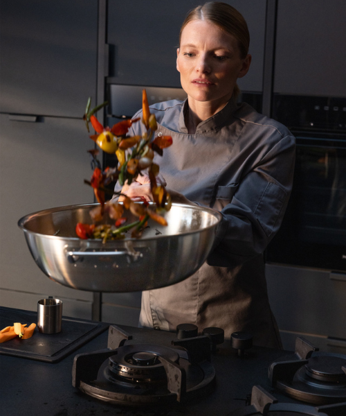 A chef in a gray uniform expertly tosses vegetables in a Fissler stainless steel pan on a simple, modern stovetop.