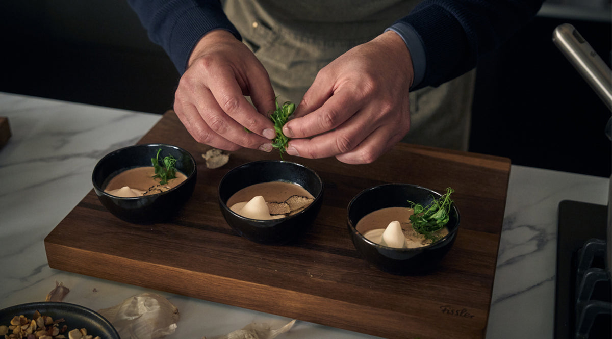 Eine Person garniert auf einem Holzbrett elegant drei kleine Schälchen mit Suppe oder Mousse mit frischen Kräutern und beweist damit ihre Liebe zum Detail. Jedes Schälchen ist mit einer runden, weißen Garnitur verziert, während daneben eine Auswahl an Zutaten kunstvoll verteilt ist. Das hochwertige Kochgeschirr und Küchenzubehör von Fissler bildet den Rahmen und unterstreicht unser Bekenntnis zu präziser Technik und zeitlosem deutschem Design.