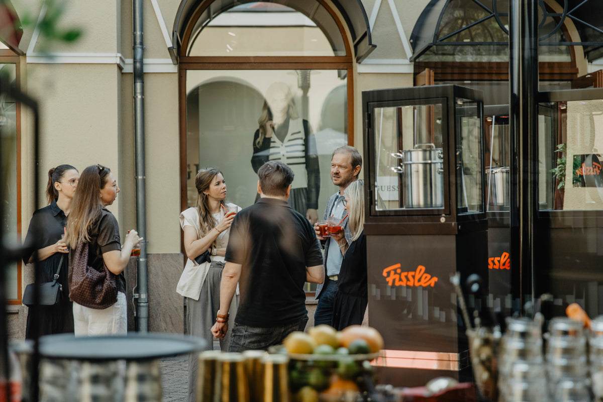 A group of people enjoy a relaxed conversation outside near a Fissler display, surrounded by our signature stainless steel cookware and kitchen accessories. Fruits are beautifully arranged on a table in the foreground beside precision-engineered pots and pans, highlighting Fissler’s timeless design and heritage of quality craftsmanship since 1845. The courtyard setting captures the casual yet refined spirit of gatherings made memorable with innovative cookware “Made in Germany.”.