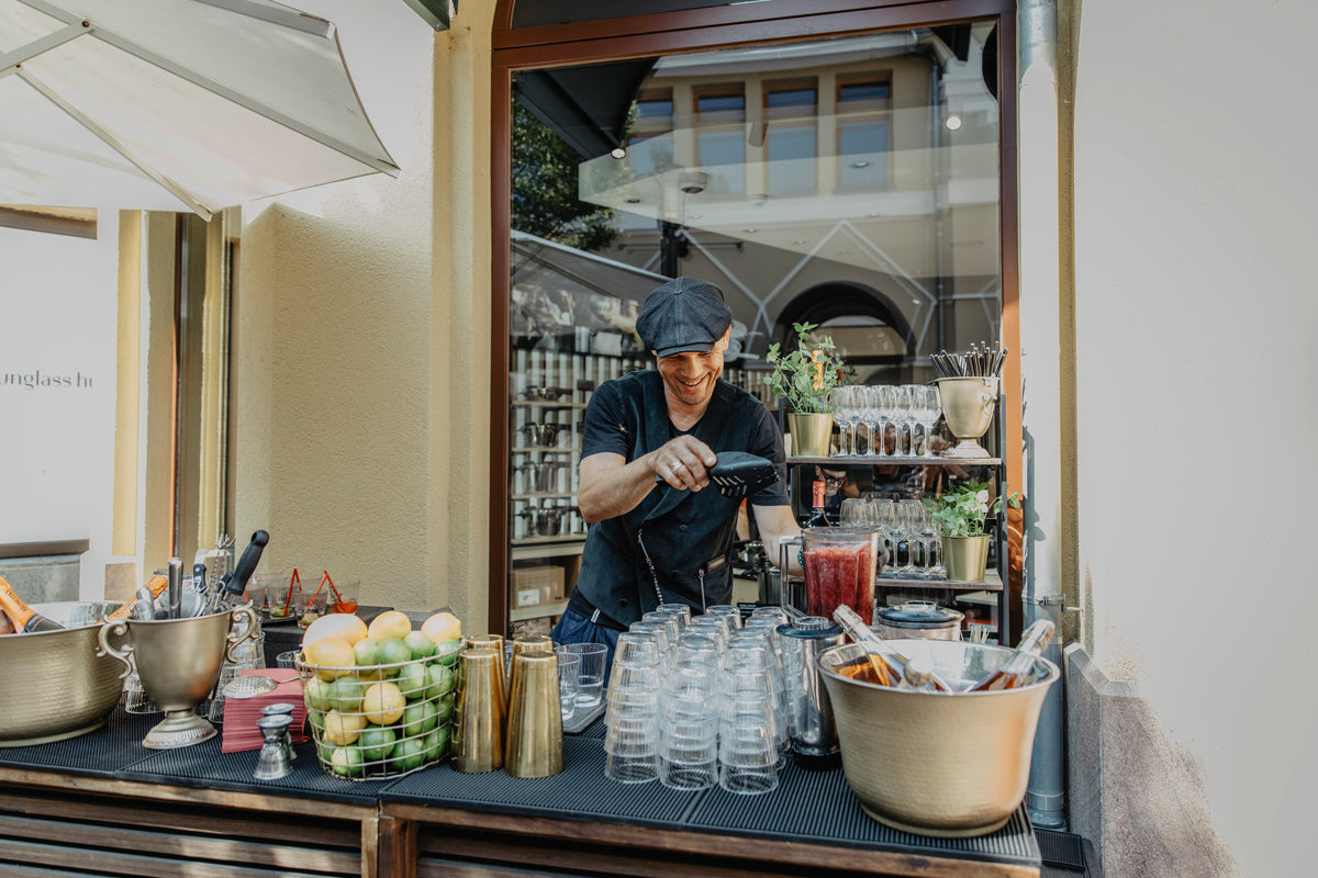 A bartender in a black uniform and hat expertly prepares drinks at an outdoor bar, surrounded by precision-engineered Fissler stainless steel barware, fresh fruit, glasses, and bottles. The refined setting reflects timeless German design and innovation, with a building elegantly visible in the background.