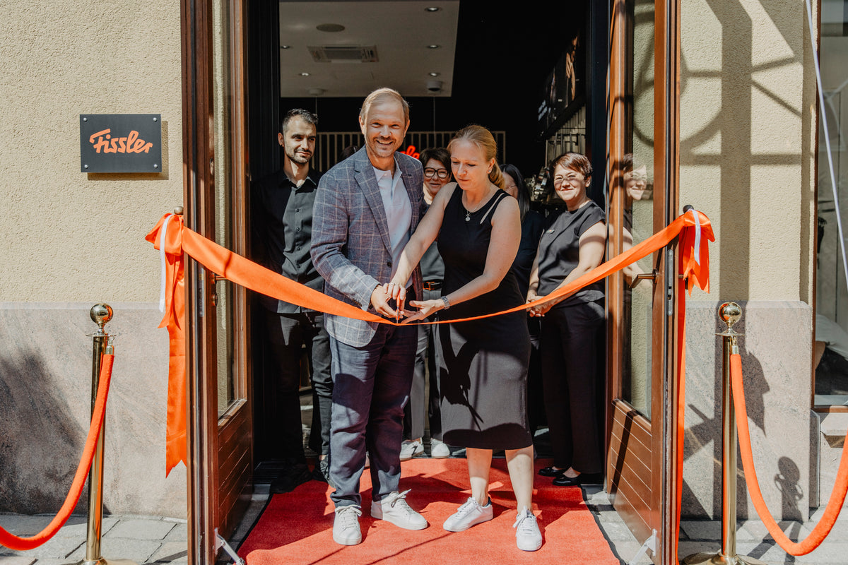 Two people perform a ribbon-cutting ceremony at the entrance of a Fissler building, with several others standing behind them on a red carpet. The Fissler sign is visible on the wall, highlighting the brand’s heritage of precision-engineered cookware made in Germany, including stainless steel pots, pans, pressure cookers, and kitchen accessories.