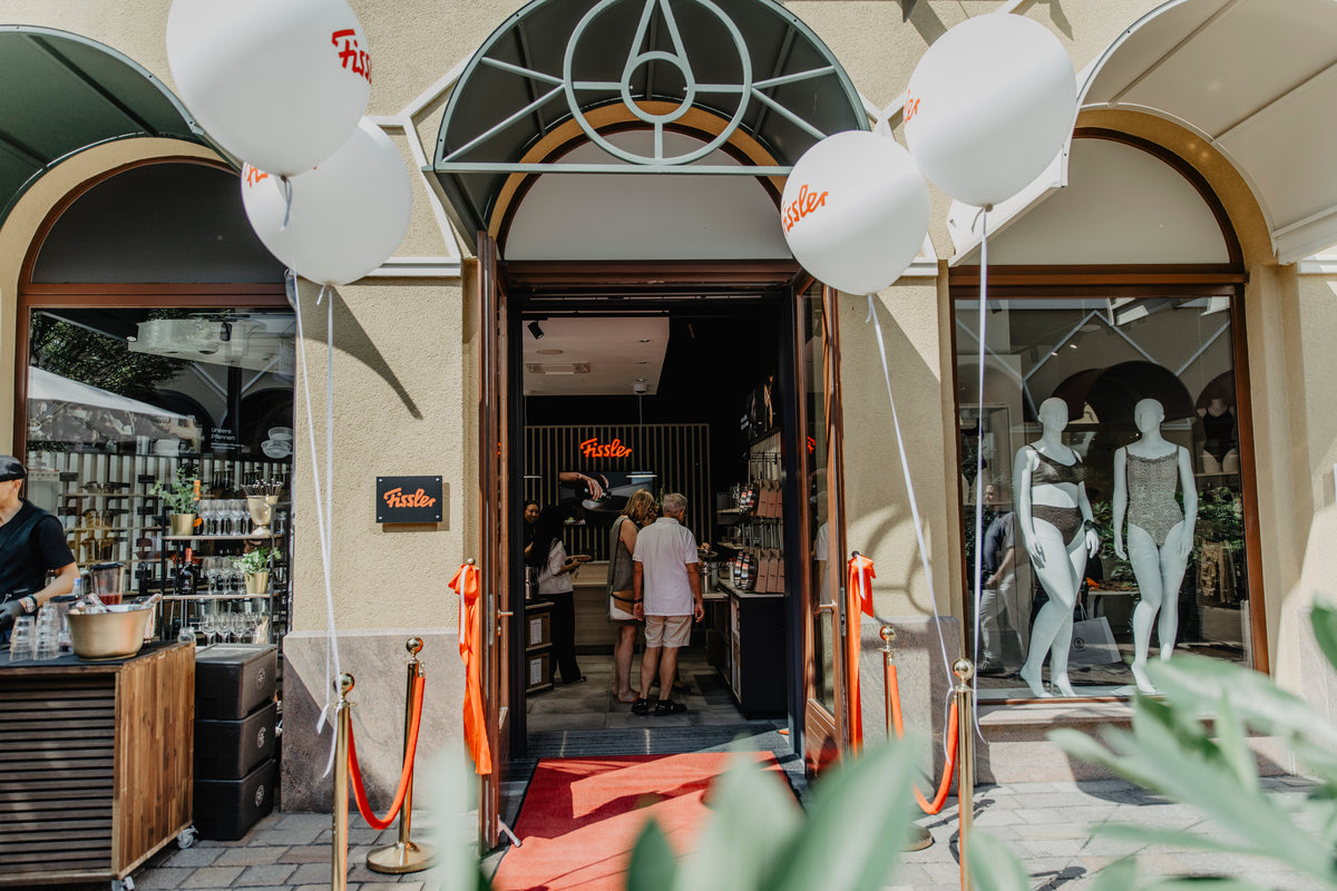 Store entrance with Fissler sign, red carpet, and white balloons. Two people enter the shop. Sleek displays of premium stainless steel cookware are visible in the window. An outdoor display on the left showcases precision-engineered pots and pans, reflecting Fissler’s heritage of quality and timeless design.