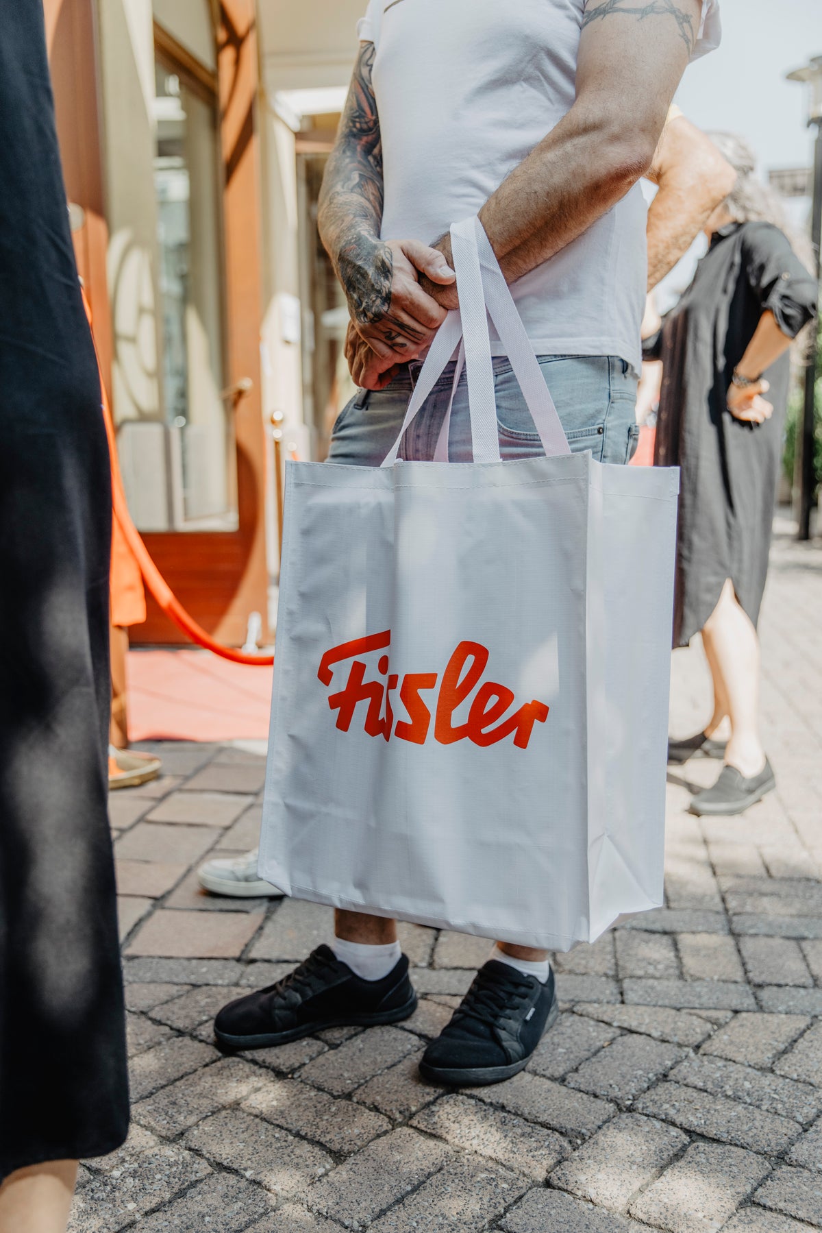A person with tattooed arms, dressed in a white t-shirt and jeans, stands on a paved walkway holding a white tote bag featuring the iconic red Fissler logo—a symbol of German precision engineering and timeless design. The background shows other people out of focus, highlighting the Fissler bag that represents our heritage of high-quality stainless steel cookware and innovative kitchen accessories.
