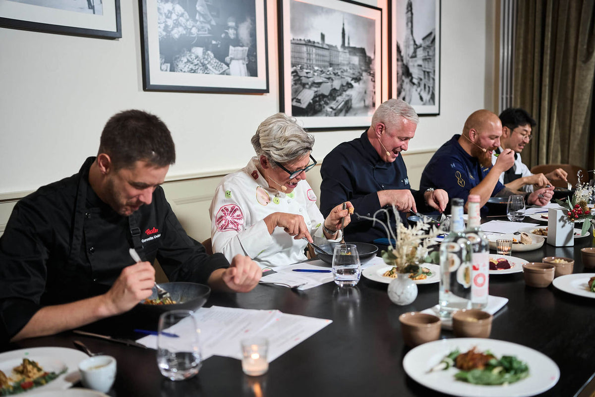 Four chefs in chef jackets judge dishes at a table set with Fissler cookware, bottles, and glasses. The wall is adorned with photos of cultural heritage.
