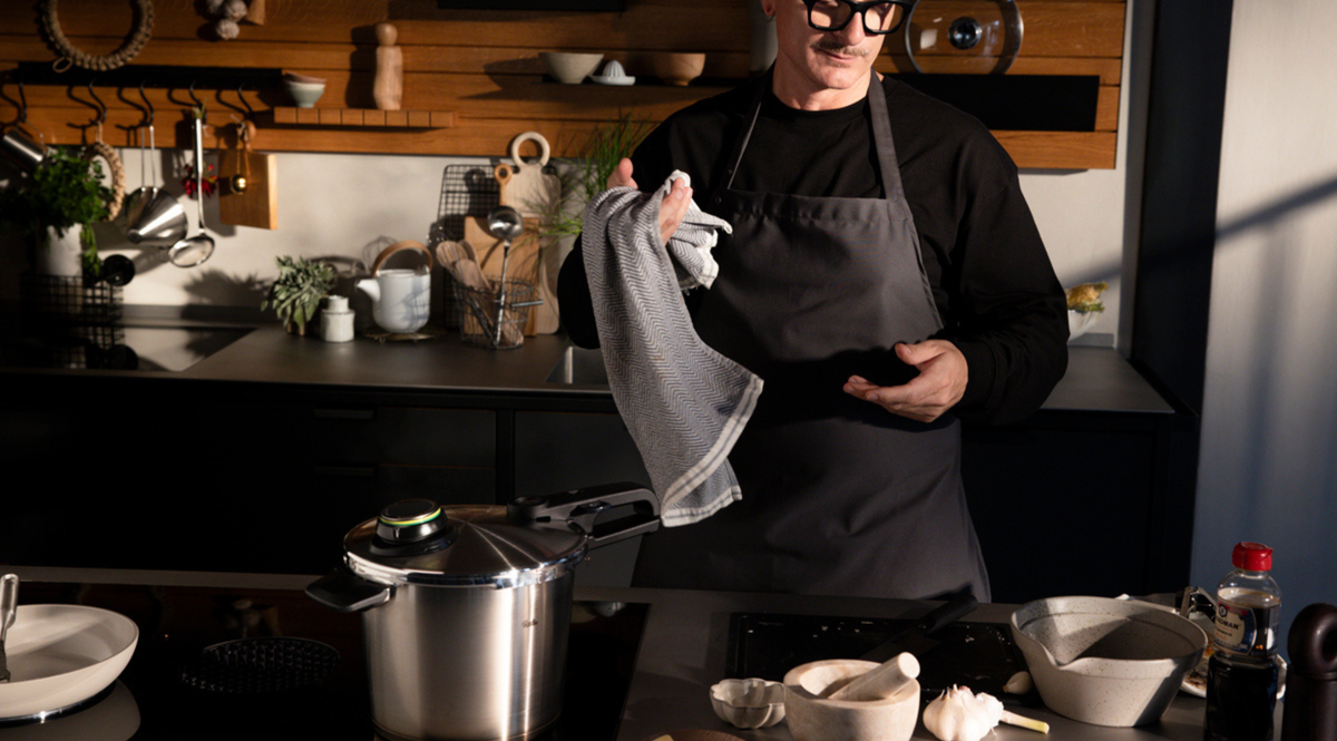 A person in a dark kitchen stands next to a Fissler pressure cooker on the stove, surrounded by premium cookware and fresh ingredients.