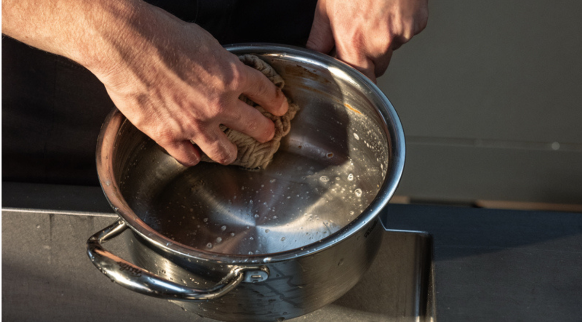 Eine Person reinigt einen Fissler-Edelstahltopf sorgfältig mit einem Schwamm und Seifenwasser über dem Spülbecken und demonstriert damit die Langlebigkeit und die beständige Qualität dieses präzise gefertigten deutschen Kochgeschirrs.
