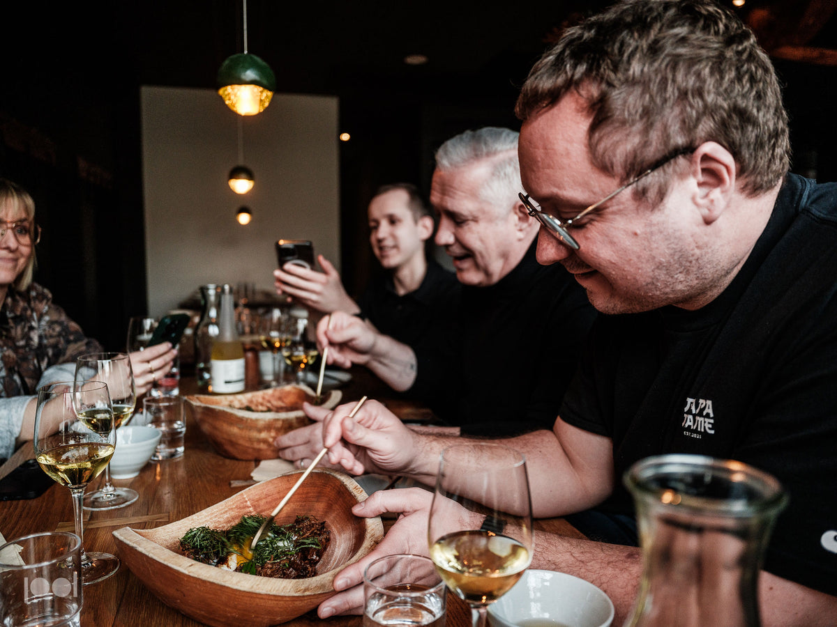 Vier Personen sitzen in einem gemütlich beleuchteten Restaurant um einen Holztisch herum und genießen gemeinsam köstliche Speisen und Getränke. Fissler-Kochgeschirr aus Edelstahl und elegantes Geschirr runden das kulinarische Erlebnis ab und spiegeln das Bekenntnis der Marke zu präziser Technik und zeitlosem deutschem Design wider. Einige Gäste lächeln, während andere Erinnerungen mit ihren Handys festhalten; Weingläser, Teller und meisterhaft gefertigte Küchenaccessoires stehen auf dem Tisch und zeugen