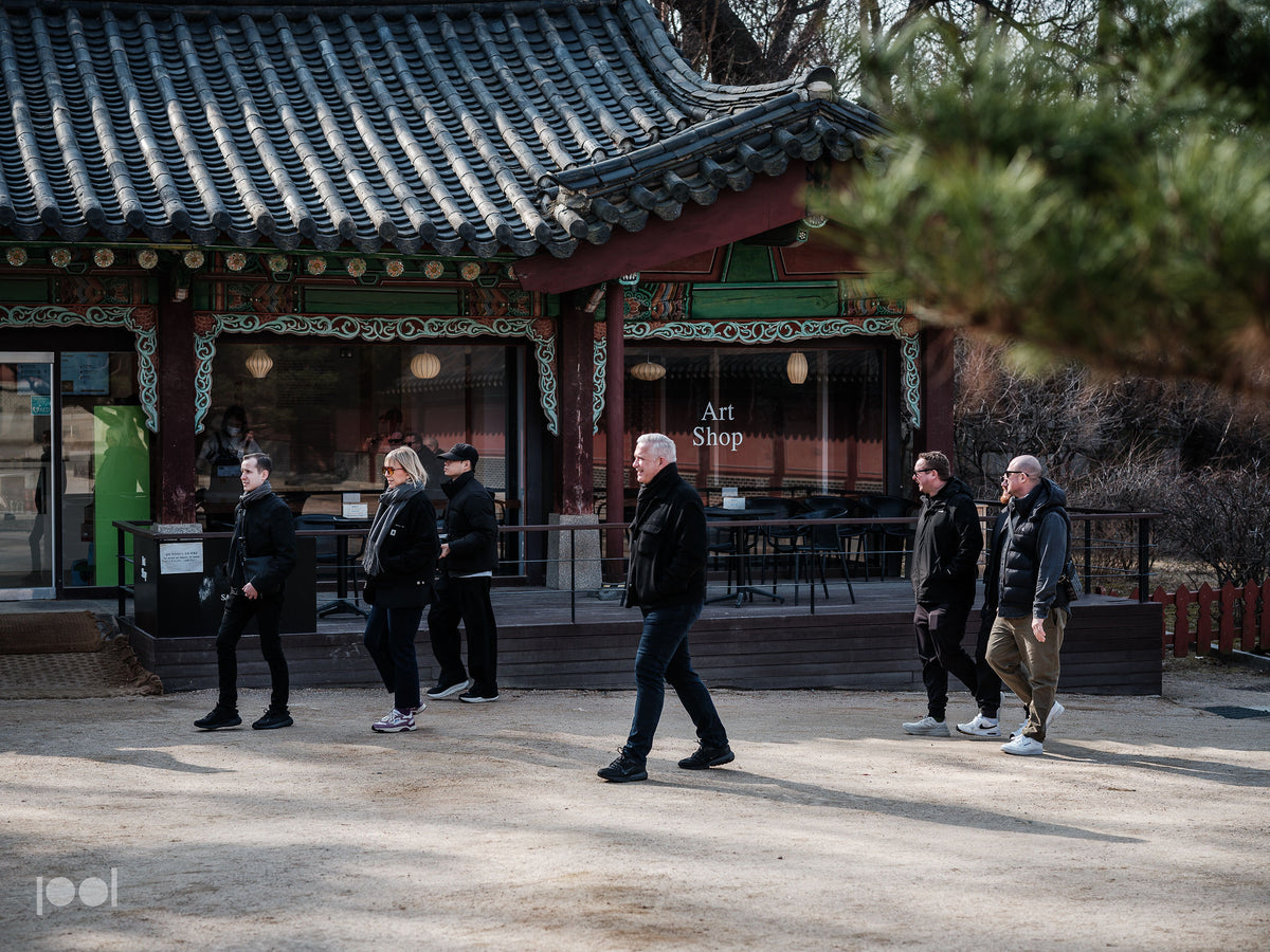 Sechs Menschen, die für kühles Wetter gekleidet sind, gehen an einem traditionellen Gebäude im koreanischen Stil vorbei, das mit kunstvollen Verzierungen und einem Ziegeldach versehen ist und an dem ein Schild mit der Aufschrift „Art Shop“ prangt. Die zeitlose Architektur und die kunstvoll gearbeiteten Details in dieser Szene spiegeln Fisslers Bekenntnis zu Qualität und Präzision wider, das sich in unseren in Deutschland hergestellten Edelstahltöpfen und -pfannen widerspiegelt. Die harmonische Verbindung
