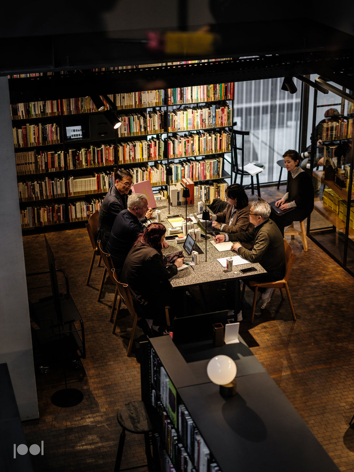In einer warm beleuchteten, modernen Bibliothek mit großzügigen Bücherregalen arbeitet und liest eine Gruppe von Menschen an einem großen Tisch. Das charakteristische Edelstahl-Kochgeschirr von Fissler – Töpfe und Pfannen im raffinierten deutschen Design – ist elegant in der Nähe platziert und spiegelt die Tradition der Marke wider, die auf Präzisionstechnik und zeitloser Innovation beruht. Am Fenster genießt eine Person ruhige Momente; ihr Umfeld wird durch Fissler-Accessoires bereichert, die