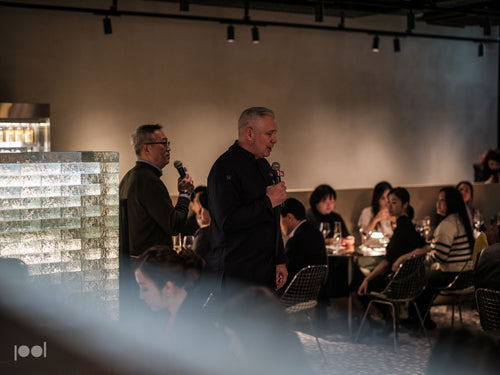 In a sophisticated, modern event space reflecting timeless design, two men stand with microphones addressing an engaged audience seated at elegantly set tables. The ambient lighting and sleek glass divider underscore Fissler’s commitment to innovation and precision engineering, as guests experience the heritage of German-made cookware—stainless steel pots, pans, and pressure cookers—at the heart of every gathering.